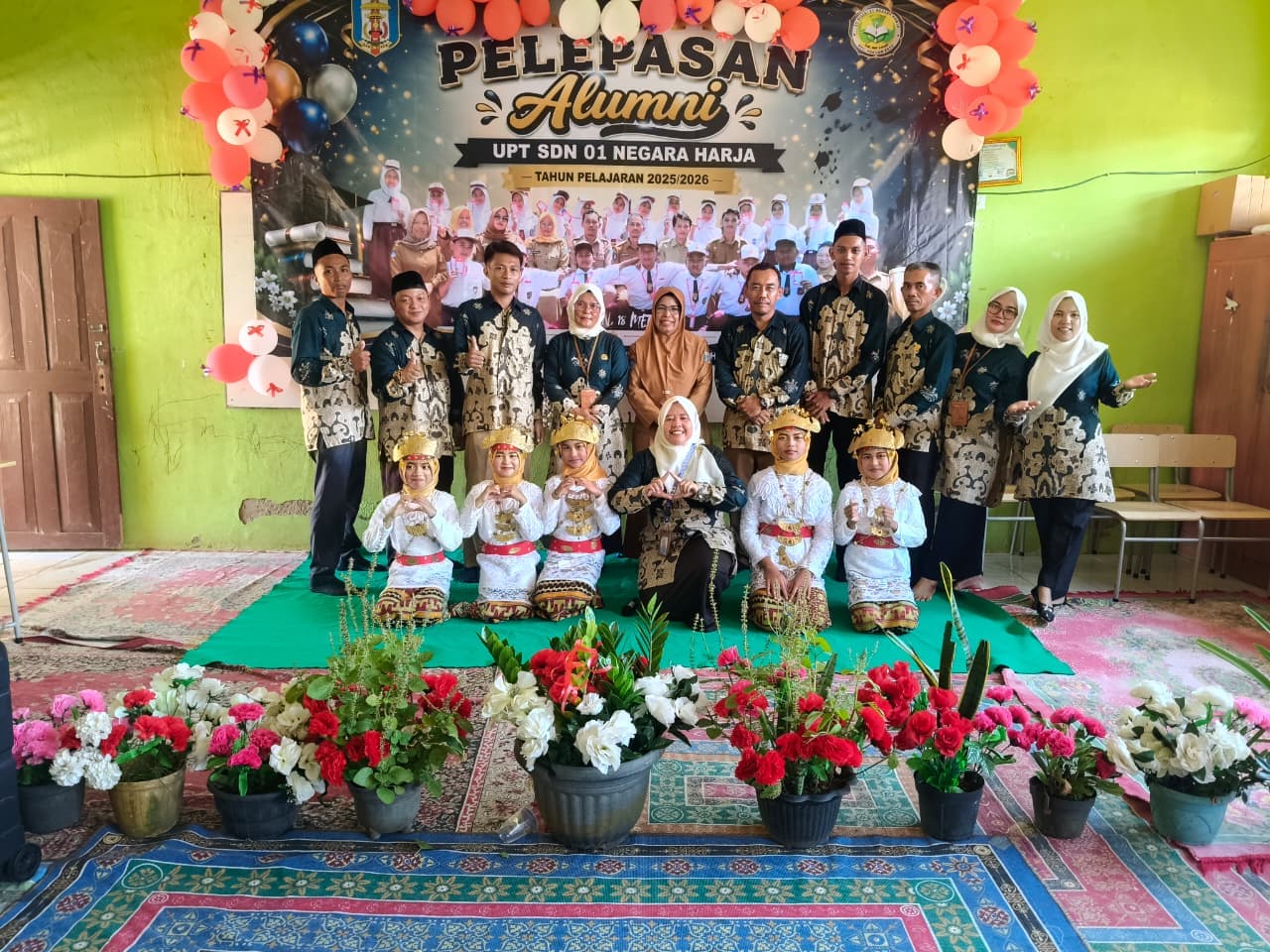 Guru berbaju batik dan penari cilik berpose di depan spanduk pelepasan alumni sekolah dasar.