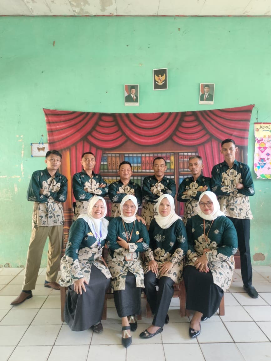 Sekelompok guru berseragam batik hijau berpose di depan latar belakang bergambar rak buku.