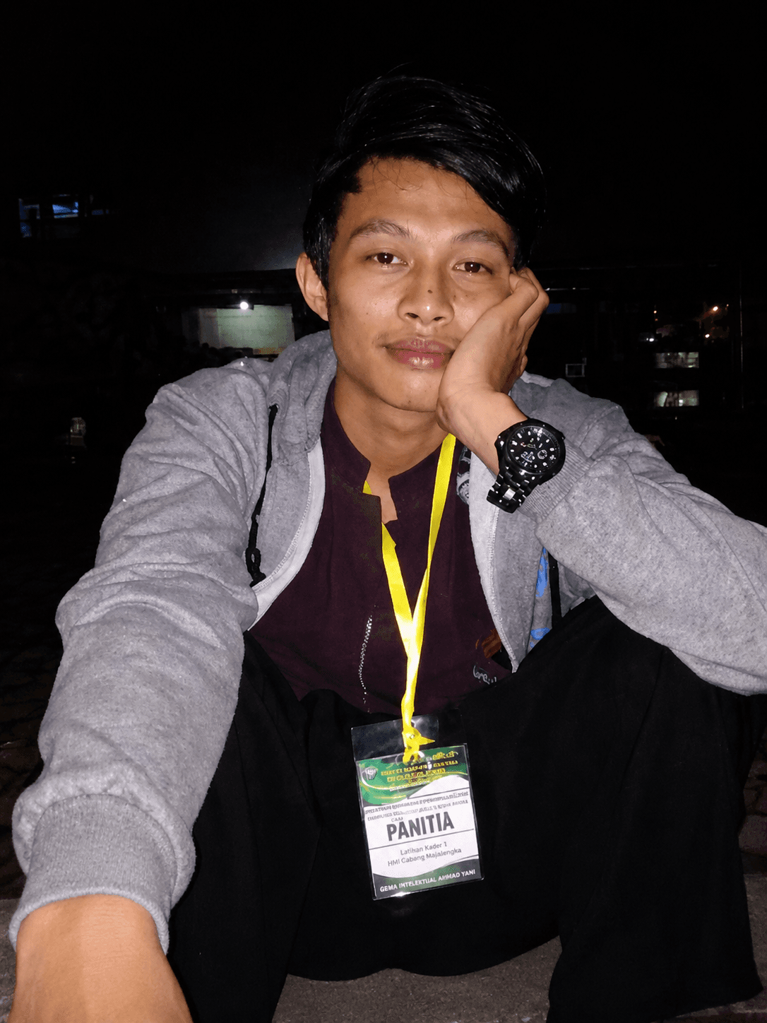 Pria muda mengenakan hoodie abu-abu dan tanda pengenal panitia, duduk santai di malam hari.
